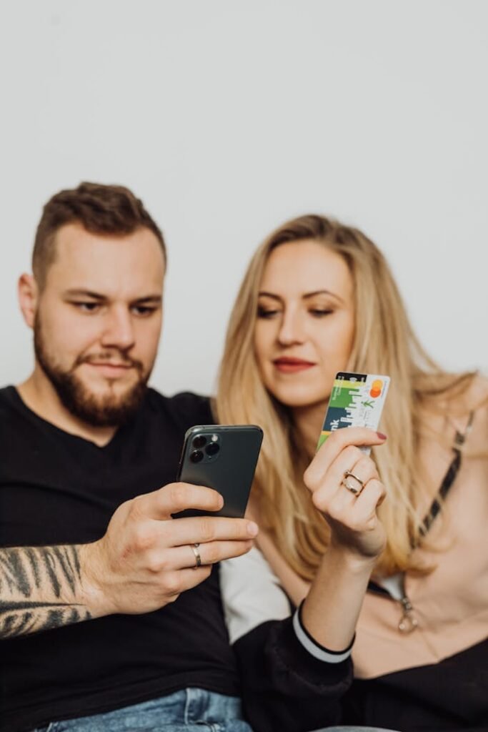 A bearded man and woman shopping online using a smartphone and credit card, sitting indoors.